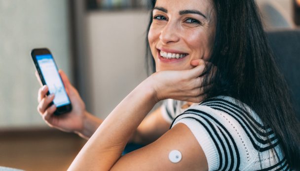 А woman smiling while holding her smartphone, with a sensor attached to her arm, likely for measuring blood sugar levels. She appears to be reviewing data or graphs from a health-related app on her phone. This scene captures the intersection of technology and personal health management, highlighting the importance of monitoring health metrics in a modern lifestyle. The woman's expression conveys positivity and engagement, making it an ideal representation of proactive health awareness.